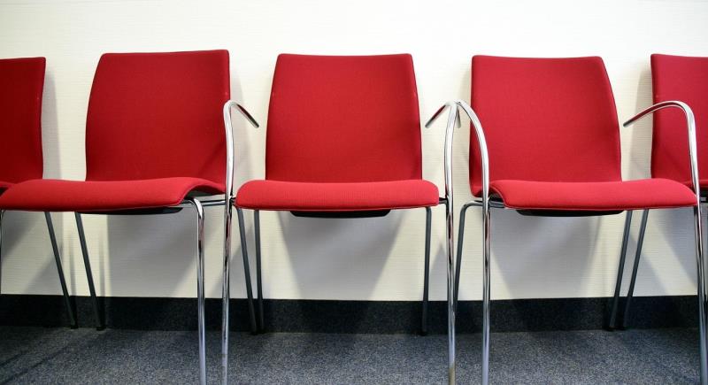 Photograph showing a row of five red chairs with metal armrests and legs against a white wall and gray carpeted floor. Chairs are evenly spaced, suggesting an office setting or waiting area. The chairs are empty. 